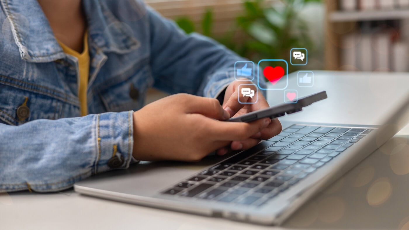 Image of a person using a smartphone while a laptop is open, with social media icons floating, such as likes, hearts, and comments, representing online interaction and digital lifestyle.
